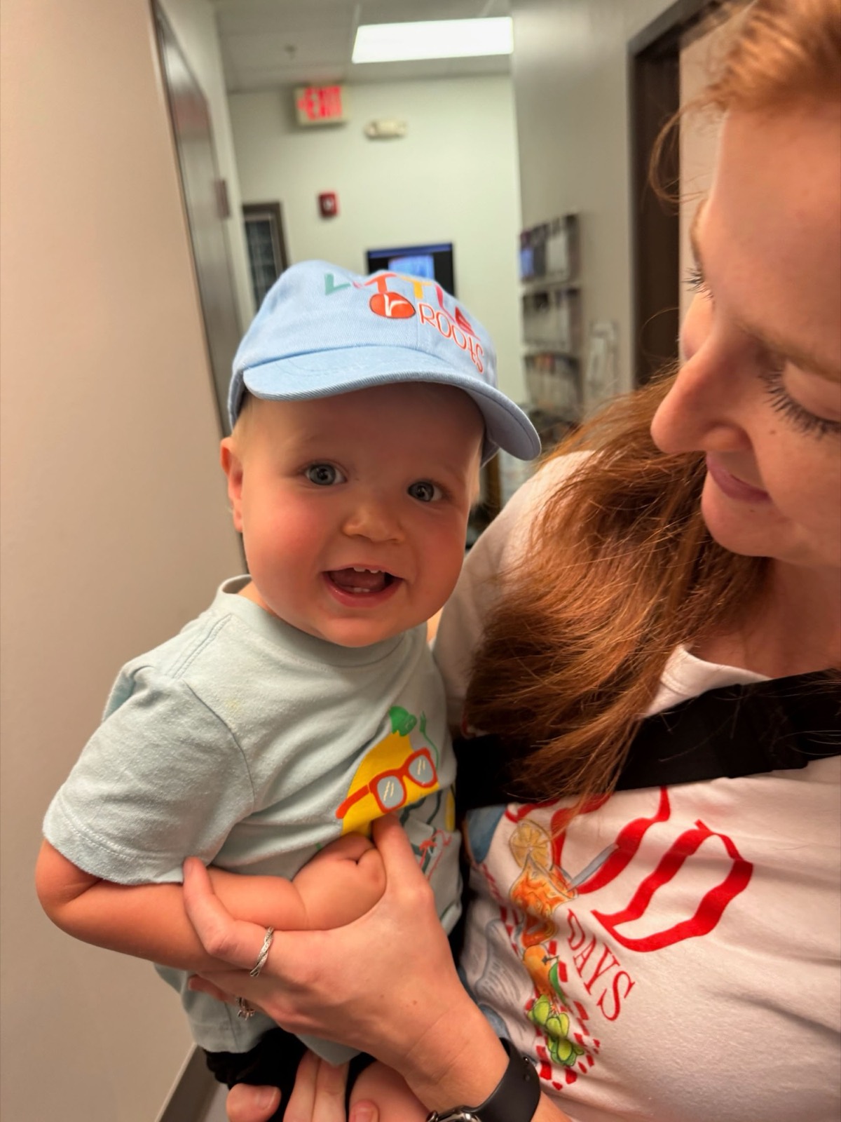 A mom holds her laughing baby wearing a Little Roots cap.