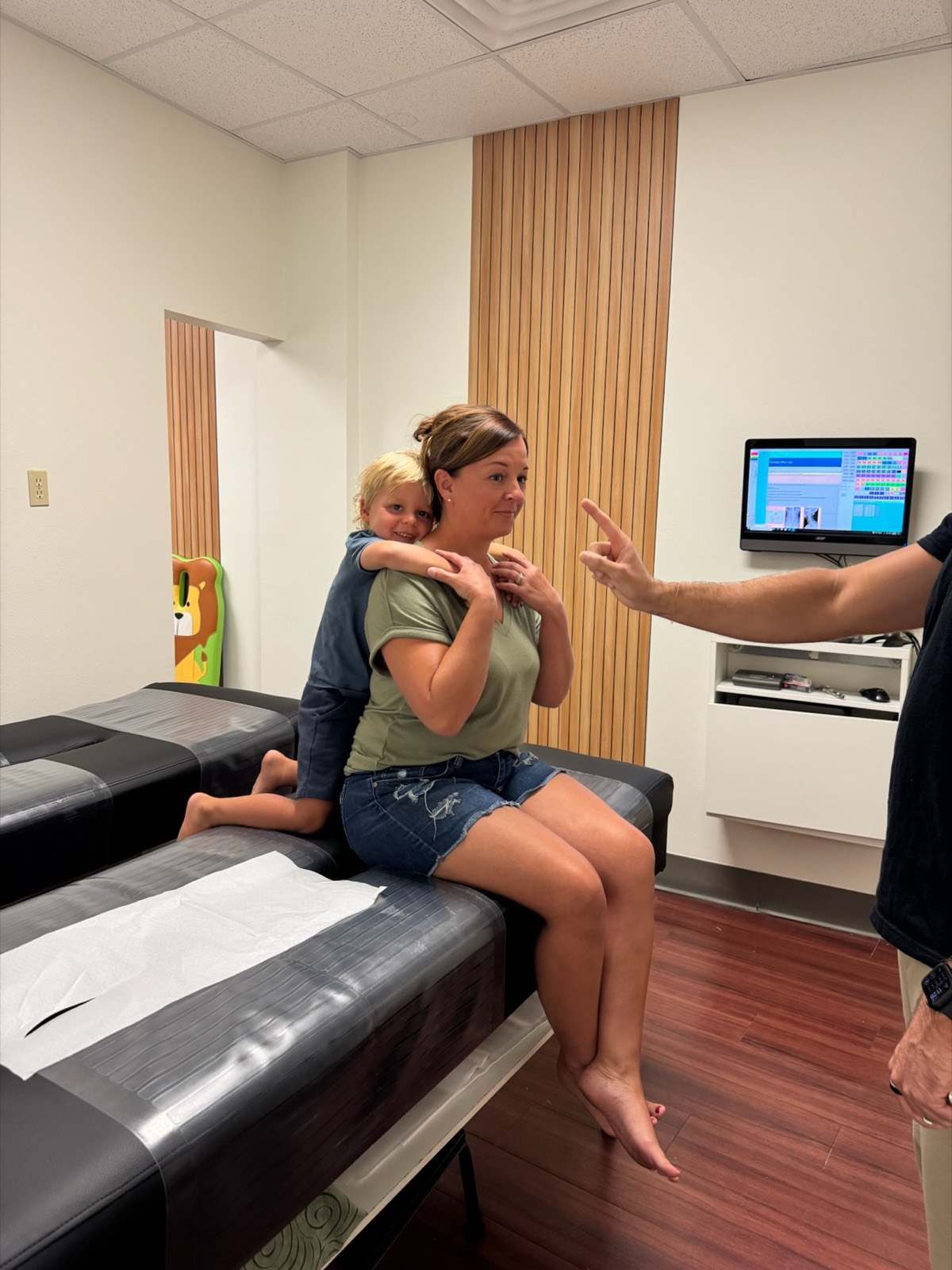 A mom sits on the adjustment table while her toddler hugs her shoulders at Little Roots.