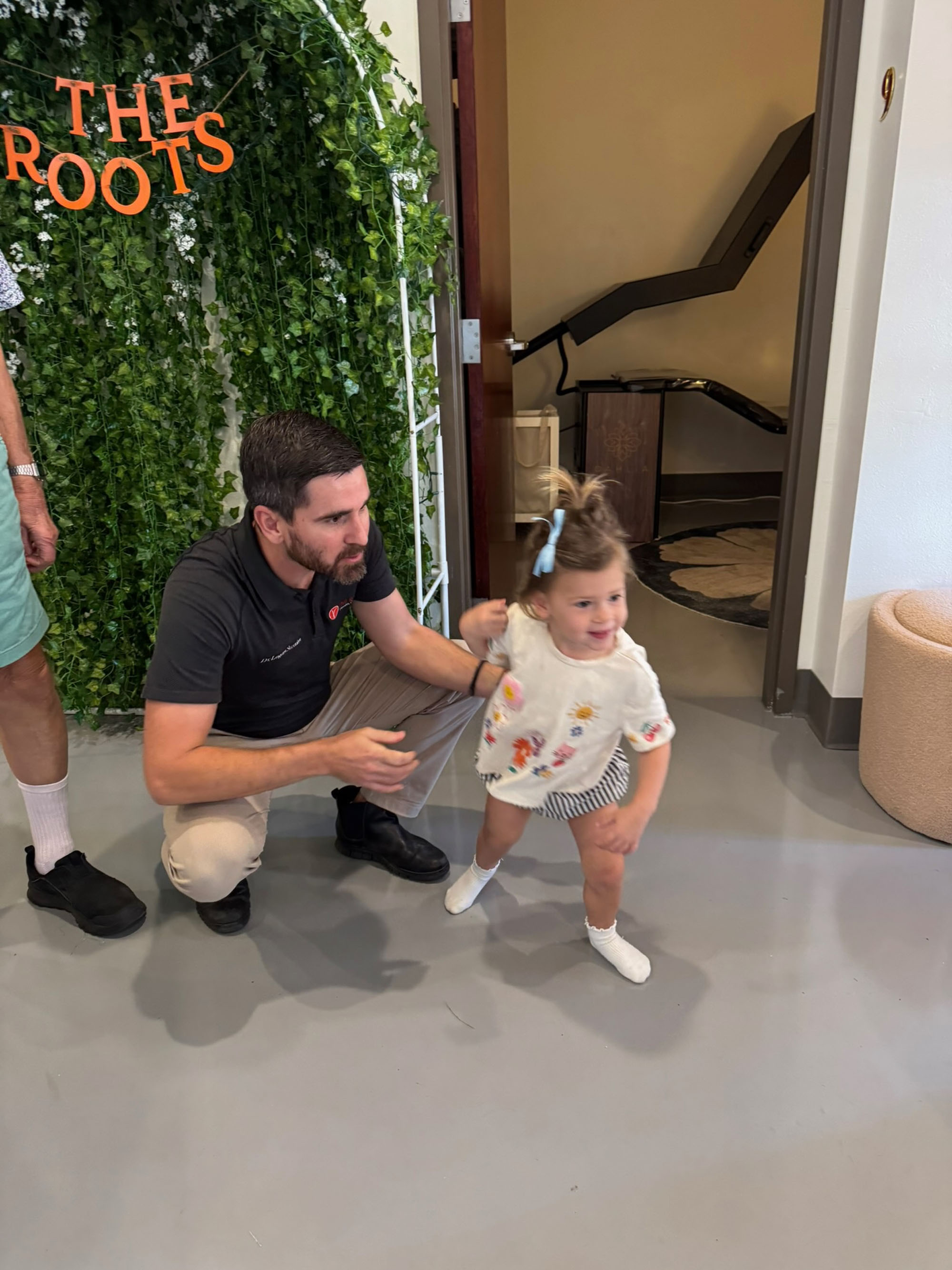 Dr. Fox kneels by The Roots arch as a toddler runs past at a family day event.